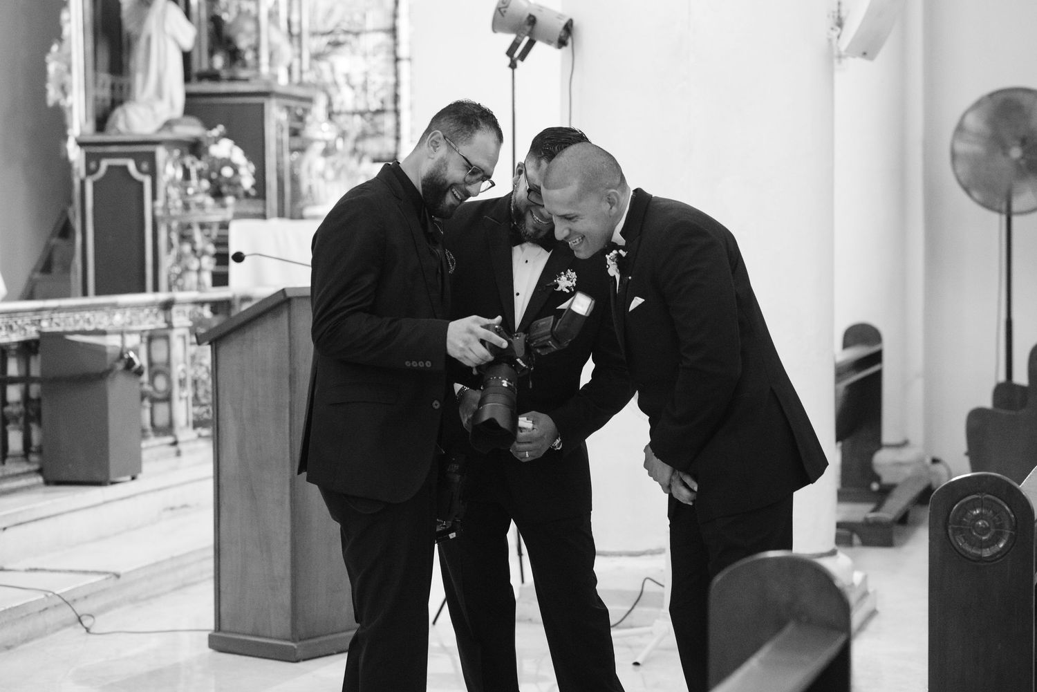 Two groomsmen in black suits assist each other with boutonnieres inside a church before a wedding ceremony.