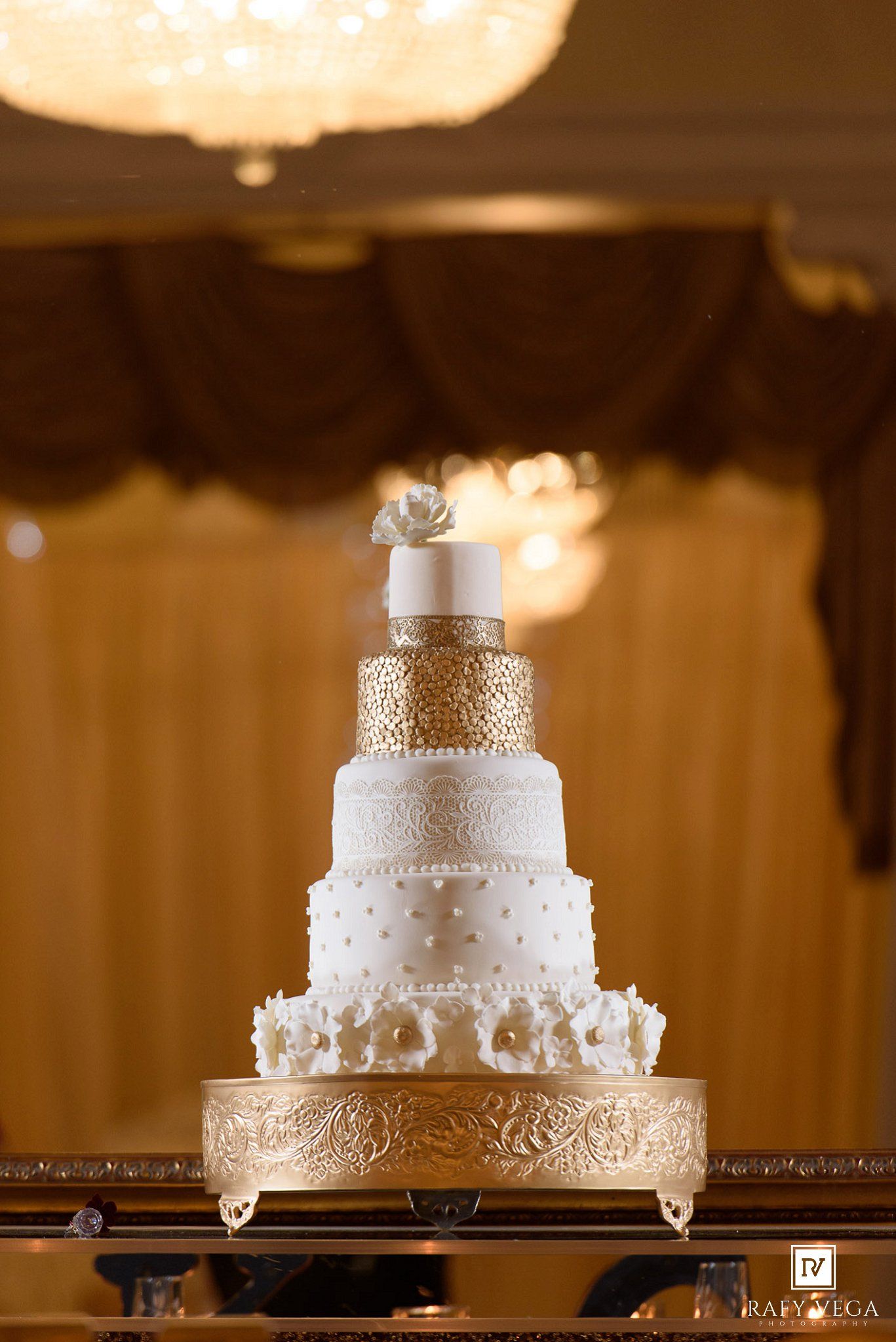 Elegant five-tier white and gold wedding cake with floral accents on a gold stand in a warmly lit reception hall.