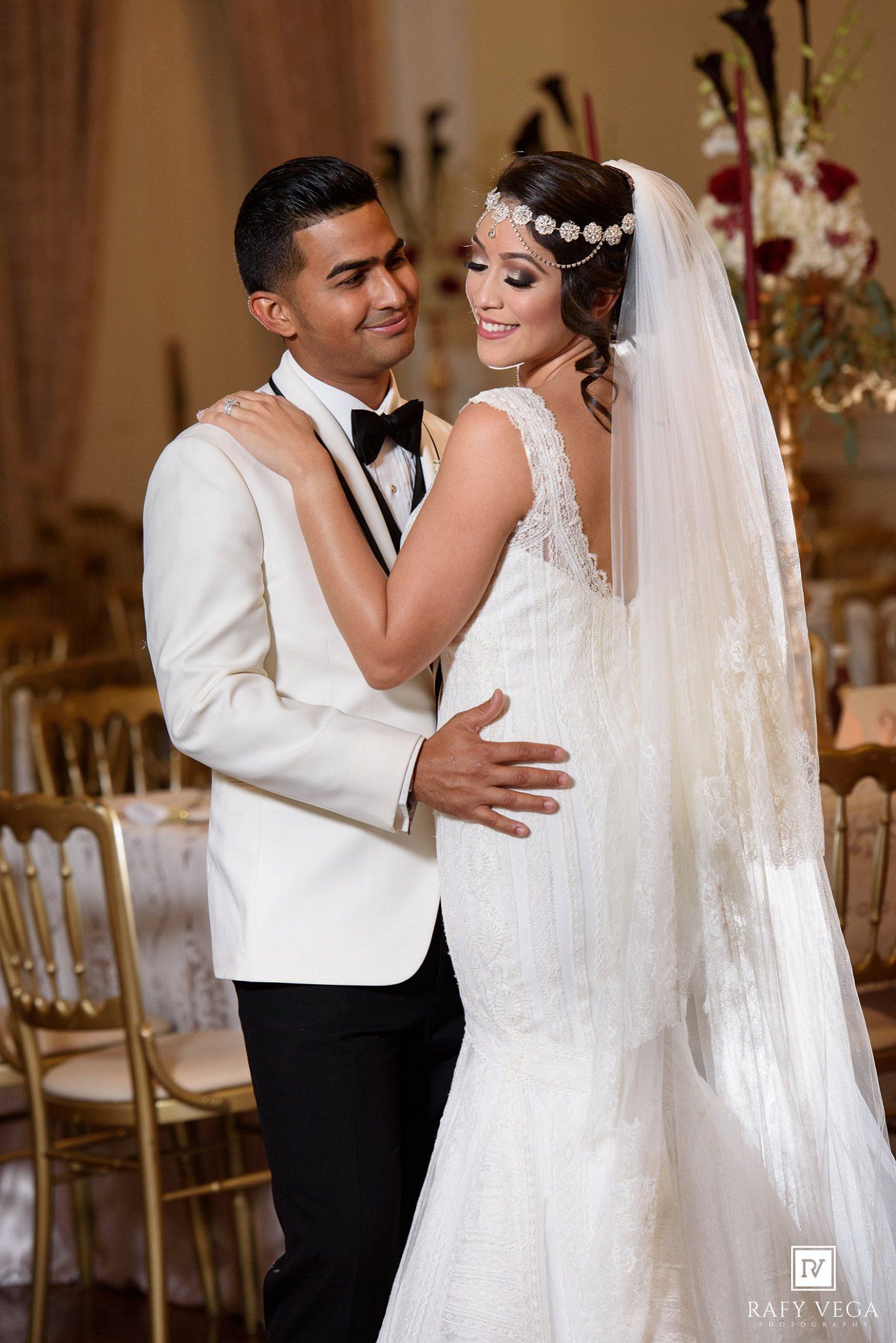 Bride and groom share first dance in elegant ballroom, bride in lace mermaid gown with veil, groom in white tuxedo.