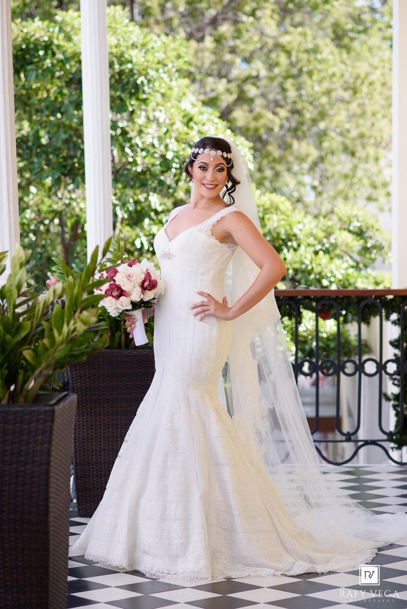 Bride in white mermaid lace wedding gown holding floral bouquet on elegant checkered floor balcony.