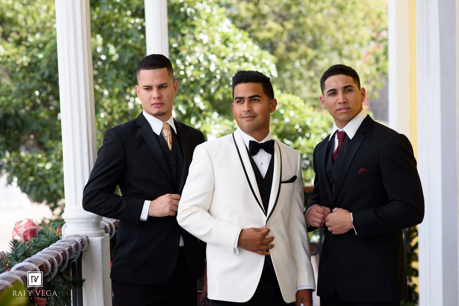 Three young men in formal black and white tuxedos pose on a elegant white columned porch for a special occasion.