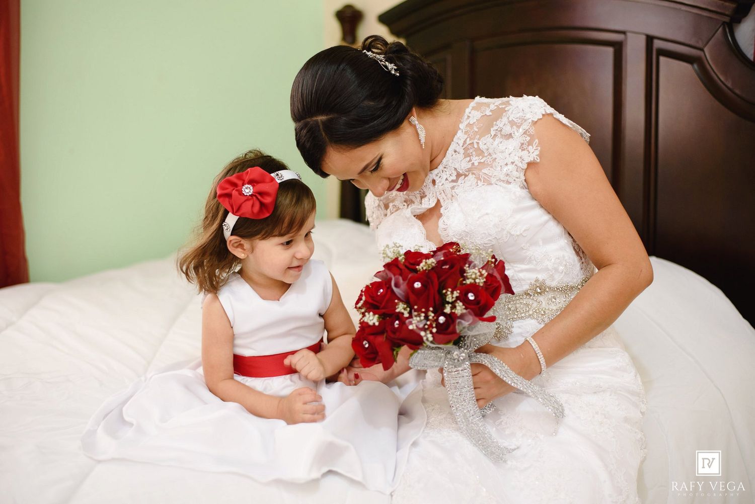 Bride in white lace gown shares bouquet of red roses with flower girl in white dress with red sash on bed.