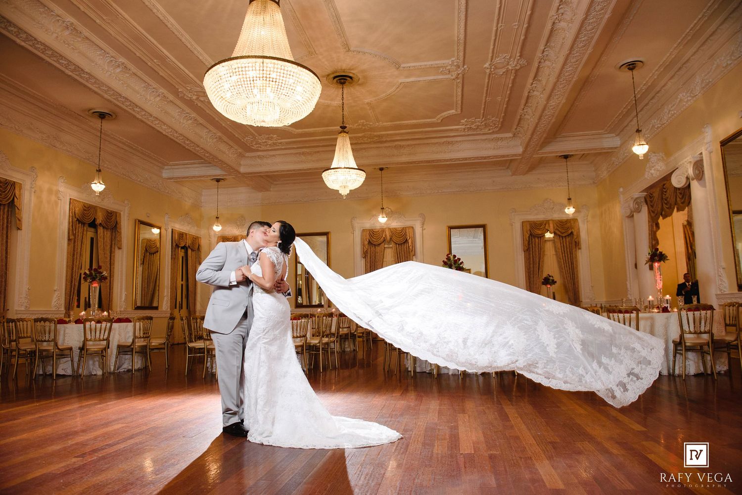 Bride and groom share a romantic kiss on dance floor, bride's veil flowing dramatically in elegant ballroom.