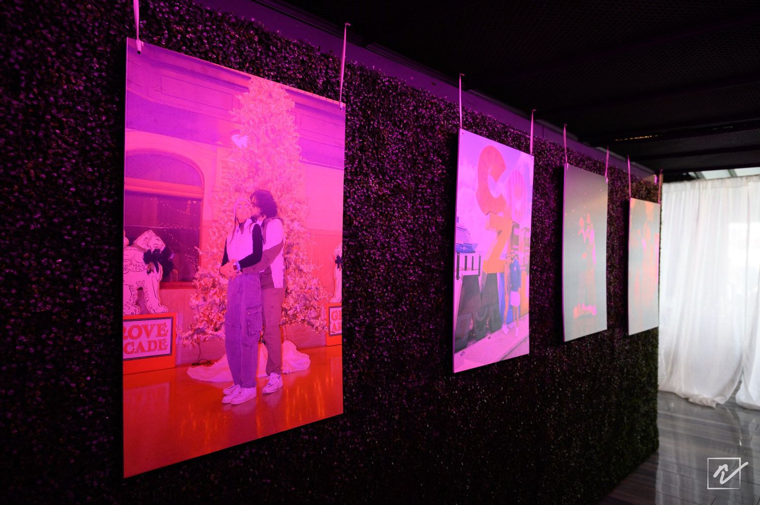 Pink-lit photo prints hang on a dark floral wall in a gallery, with one showing two people dancing by a white Christmas tree.