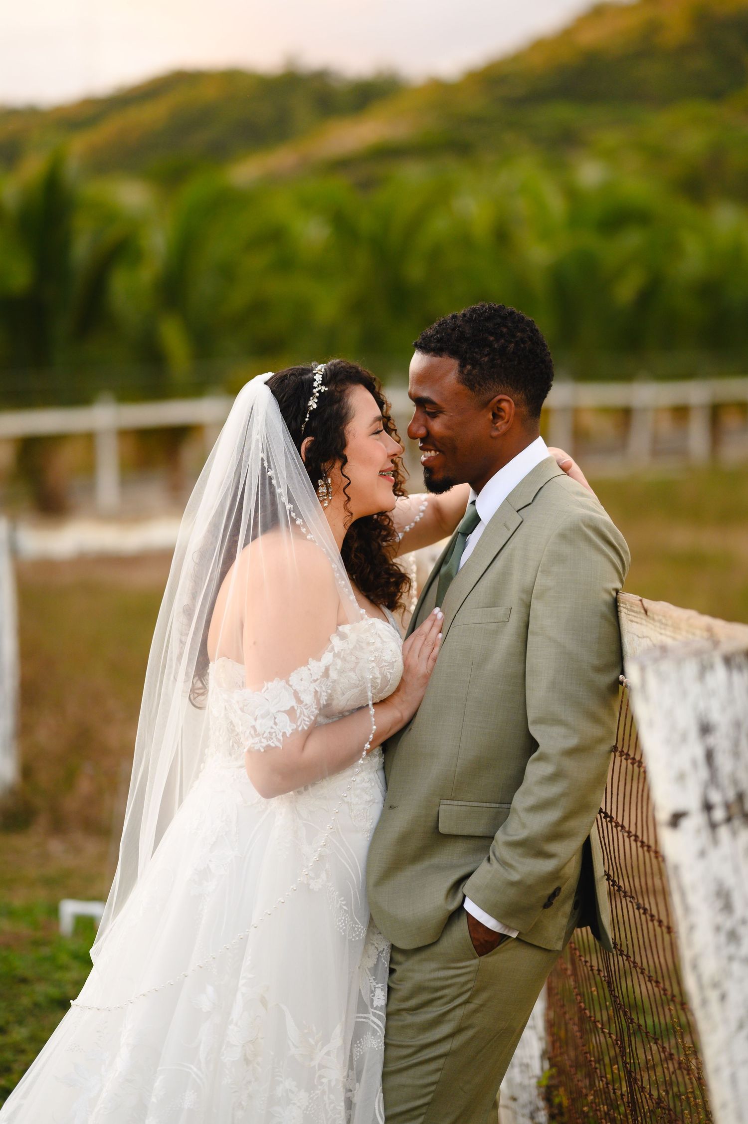 Bride in a lace gown and veil embraces her groom in a sage green suit beside a rustic fence with lush green hills in the background.