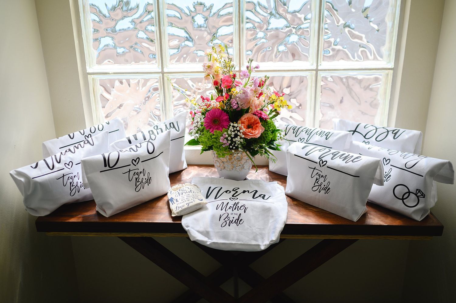 Personalized white bridal party cosmetic bags with black script lettering displayed on a wooden table by a window.