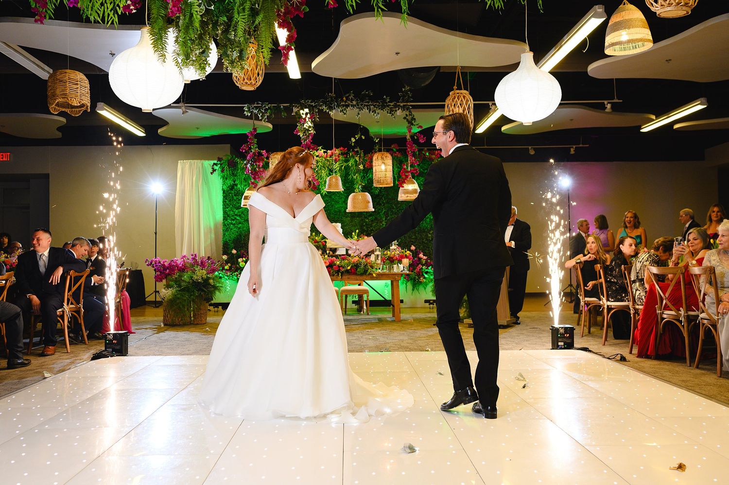 Bride and groom share first dance on white floor surrounded by sparkling fountains and hanging lanterns at wedding reception.