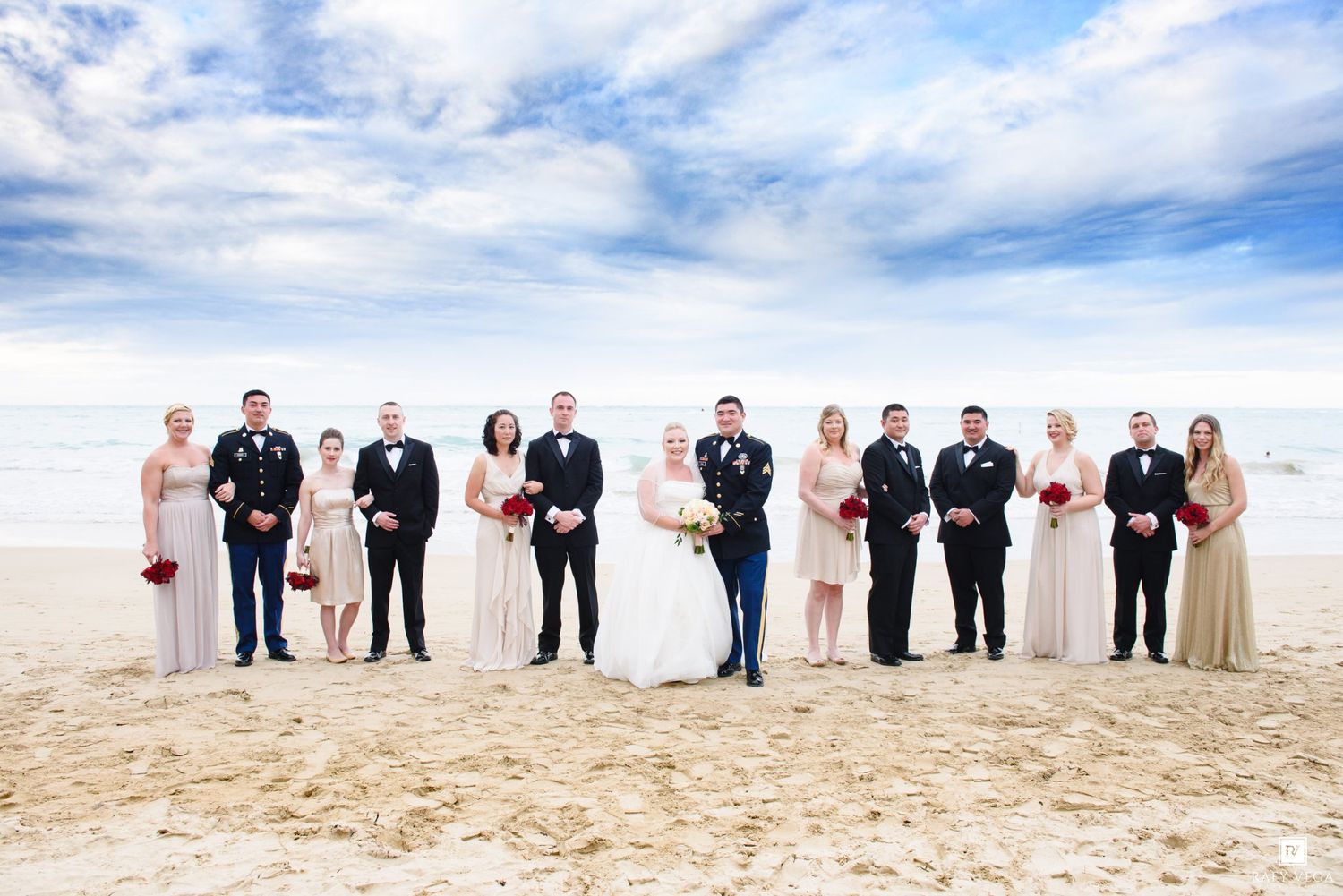 Wedding party poses on sandy beach with blue sky, bride and groom centered among groomsmen and bridesmaids in formal attire.