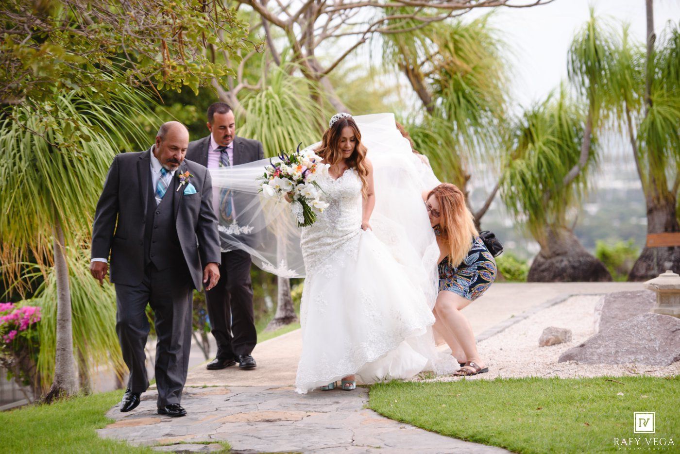 Bride in white gown walks with escorts as assistant adjusts veil outdoors at elegant garden wedding venue.