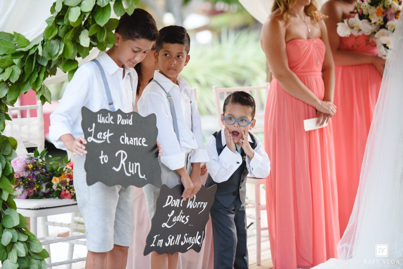 Three young ring bearers hold humorous chalkboard signs at an outdoor wedding ceremony with bridesmaids in coral dresses.