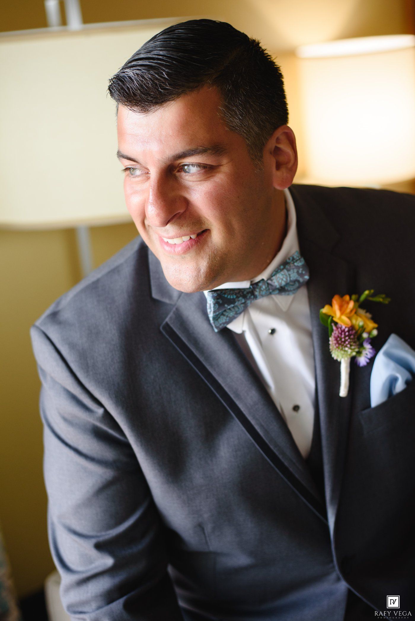 Smiling groom in gray tuxedo with floral boutonniere and teal bow tie on wedding day.