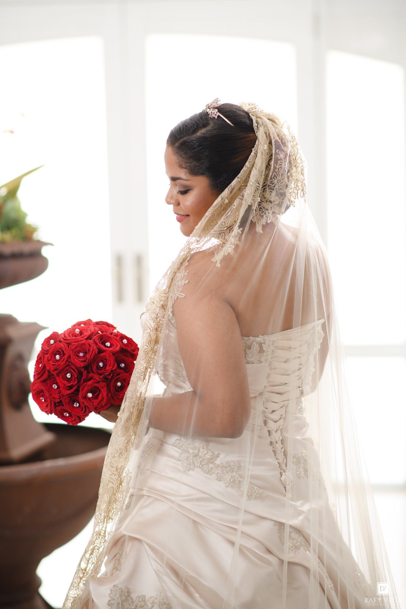 Bride in white lace wedding gown holding red rose bouquet, wearing gold-trimmed veil, viewed from behind.