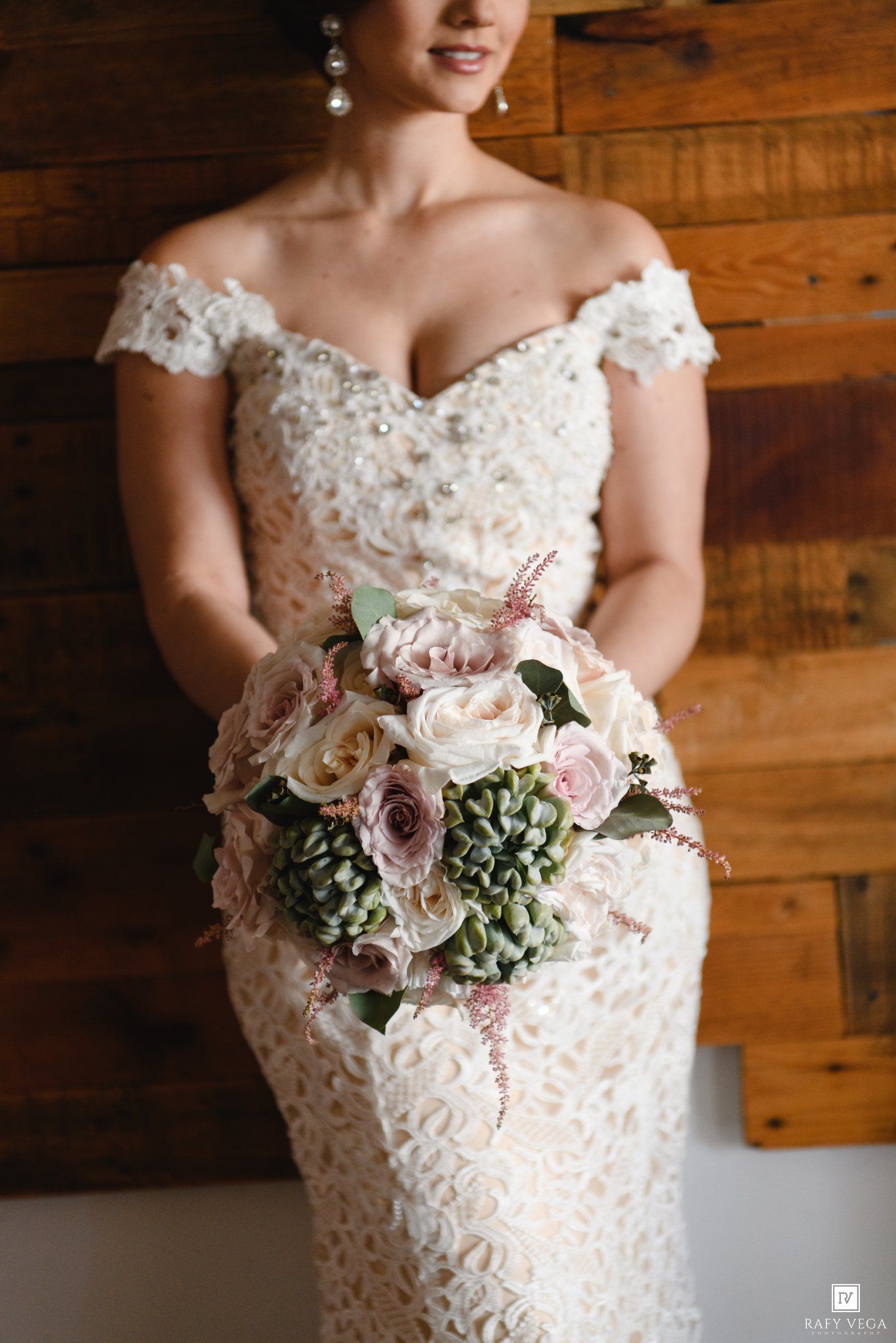 Bride in lace off-shoulder wedding gown holding blush pink and green bouquet of roses against rustic wood wall.