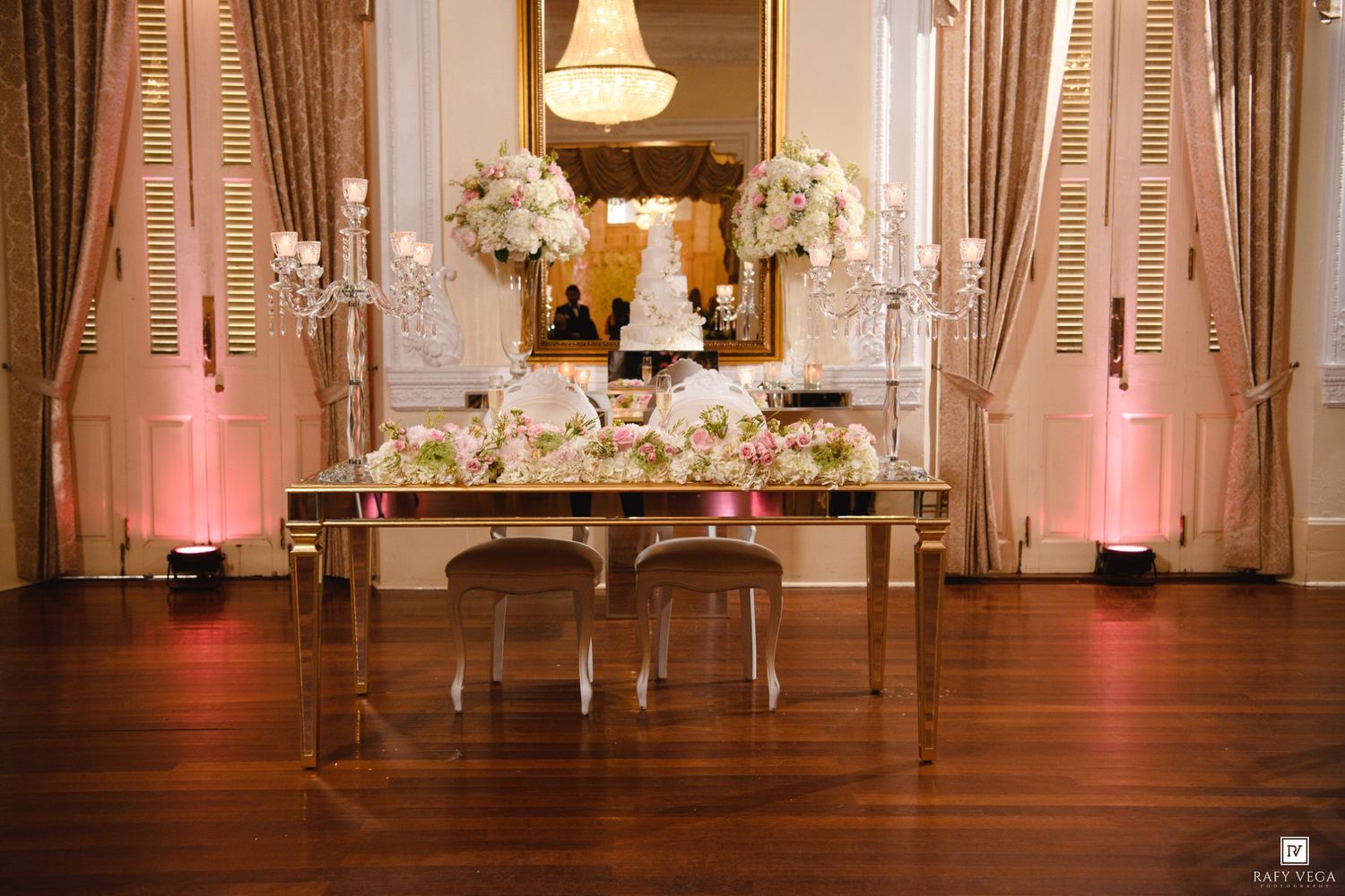 Elegant wedding sweetheart table with floral arrangements, candelabras, and tiered cake in a grand ballroom.