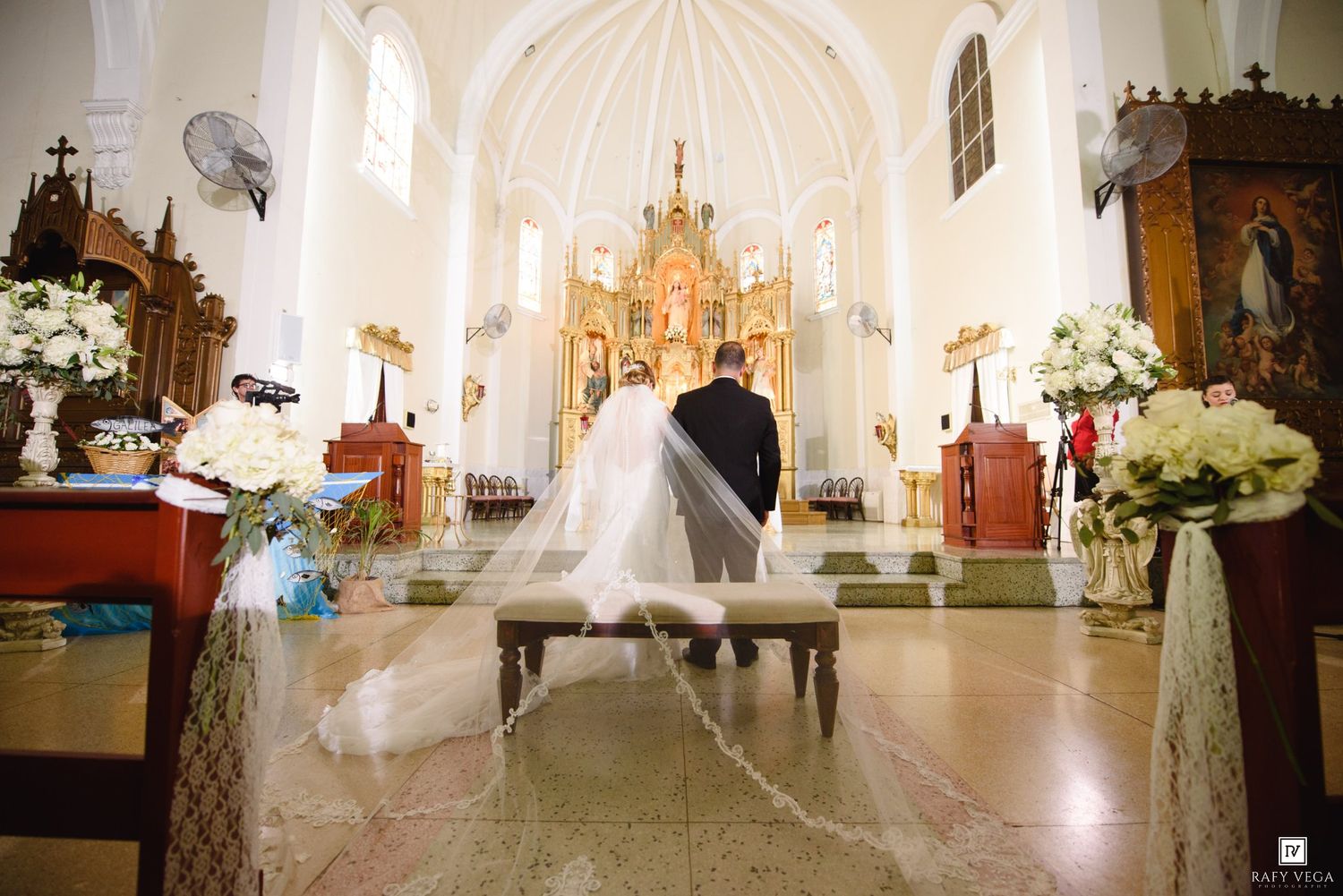 Bride and groom walk down the aisle of an elegant white church decorated with floral arrangements during a wedding ceremony.
