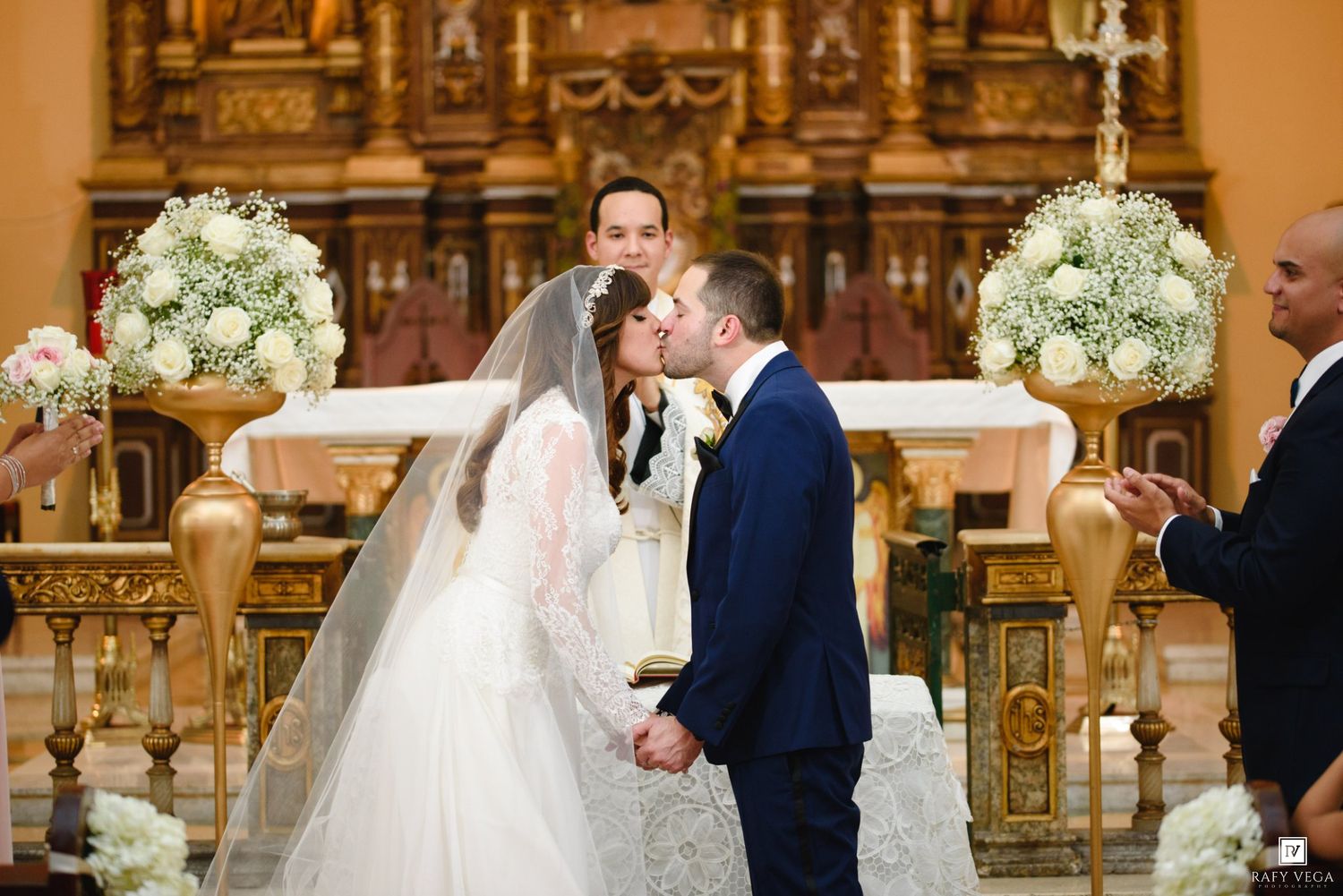 Bride and groom share first kiss at ornate church altar adorned with white floral arrangements during wedding ceremony.