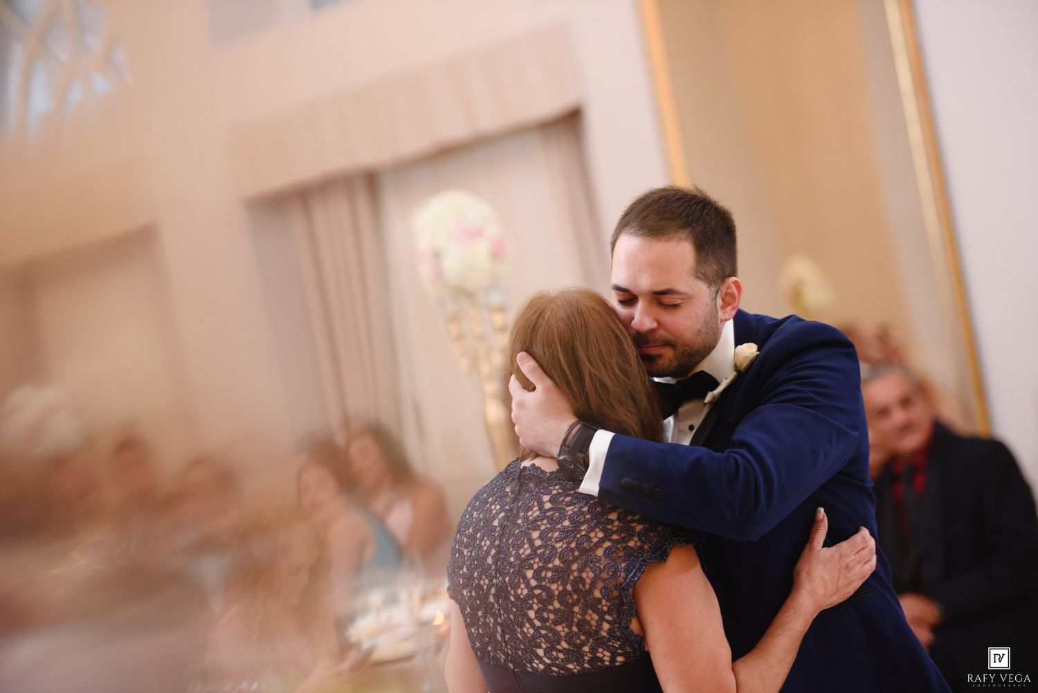 Groom in navy suit shares emotional embrace with guest during wedding reception dance.