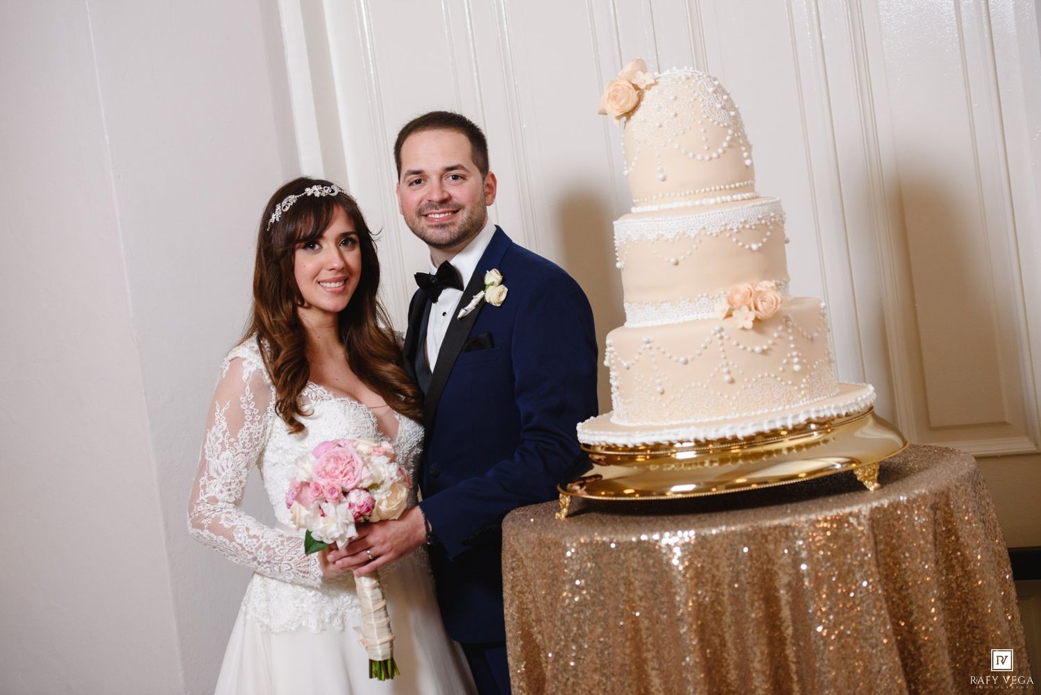 Bride and groom smiling beside elegant four-tier white wedding cake on gold sequin tablecloth.