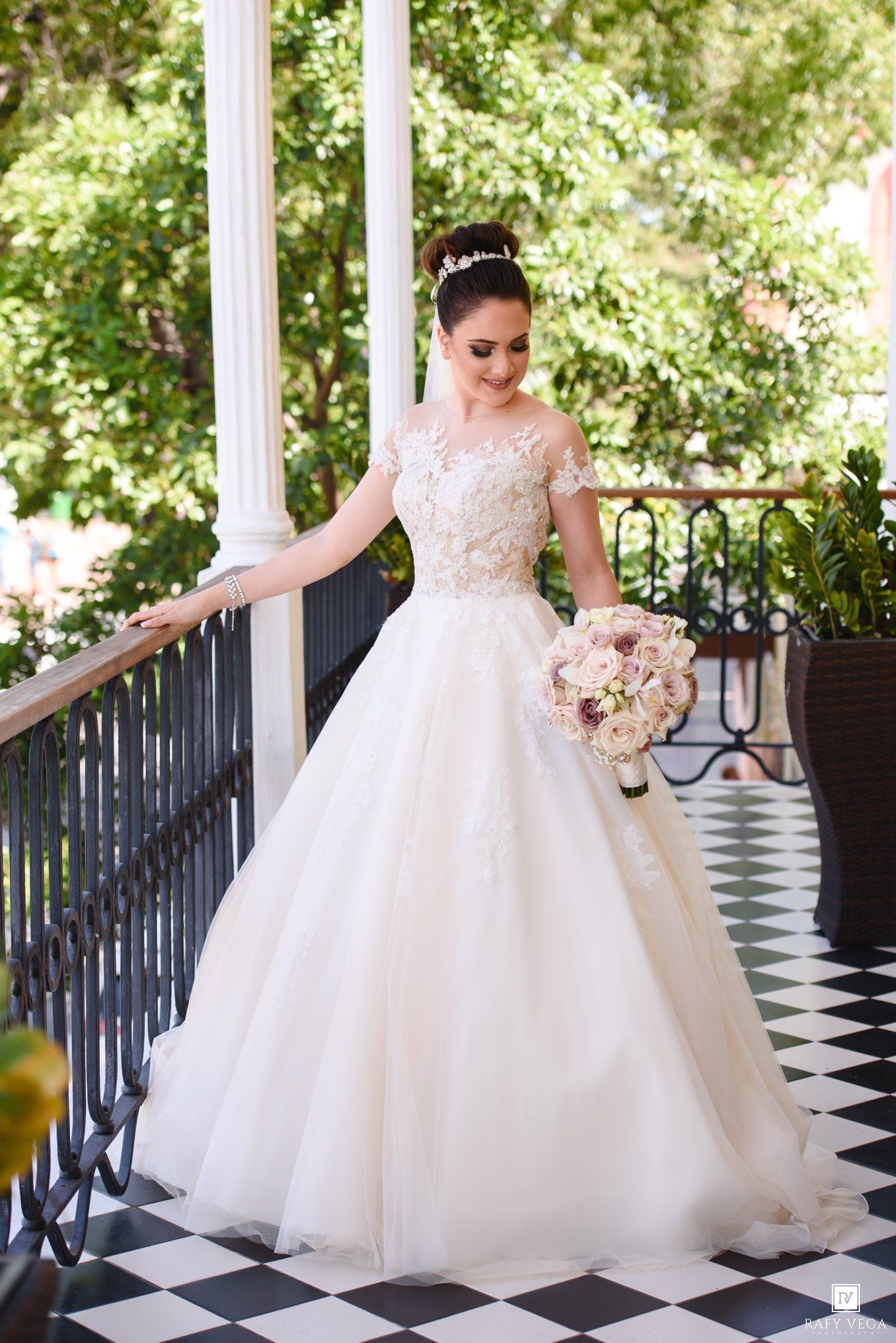 Bride in a white lace ballgown wedding dress holding a bouquet on a balcony with checkered floors.