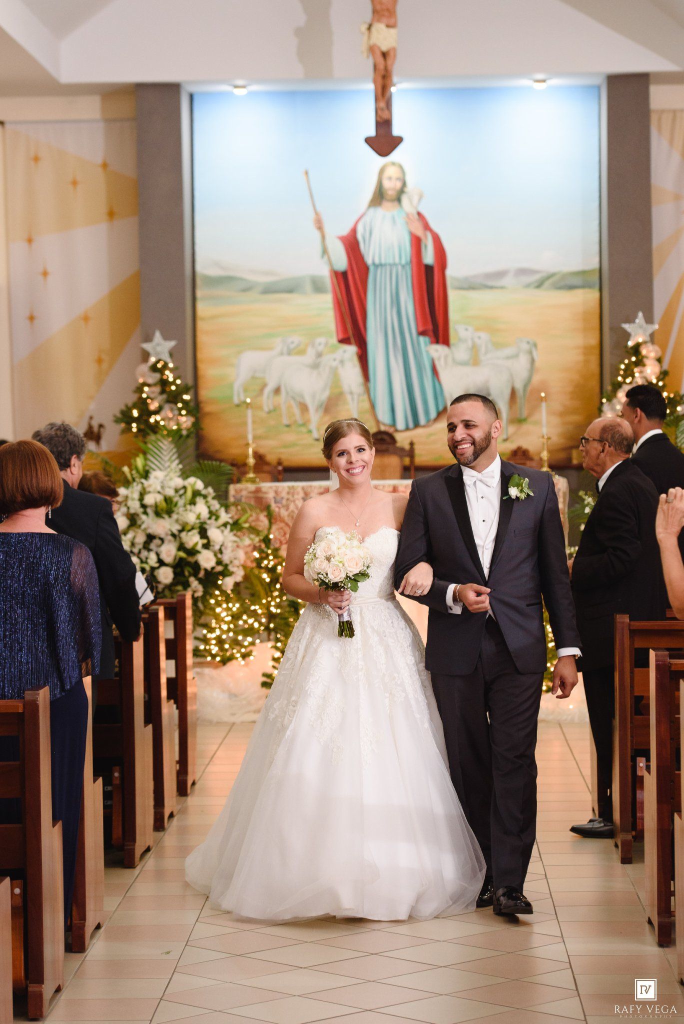 Bride and groom walk down church aisle after wedding ceremony, with Jesus painting backdrop and floral arrangements.