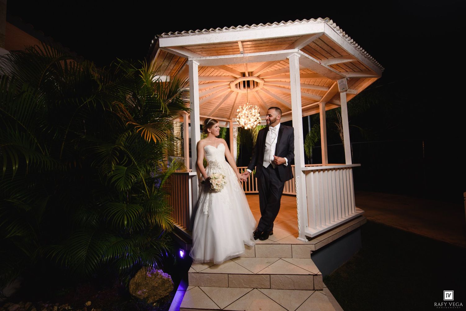 Bride and groom exit a lit gazebo at night, surrounded by lush tropical greenery at their wedding venue.