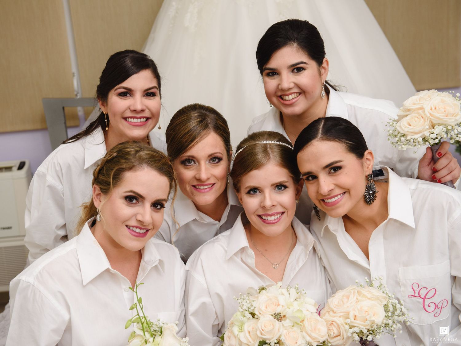 Six bridesmaids in white button-up shirts pose together holding white bouquets before a wedding ceremony.