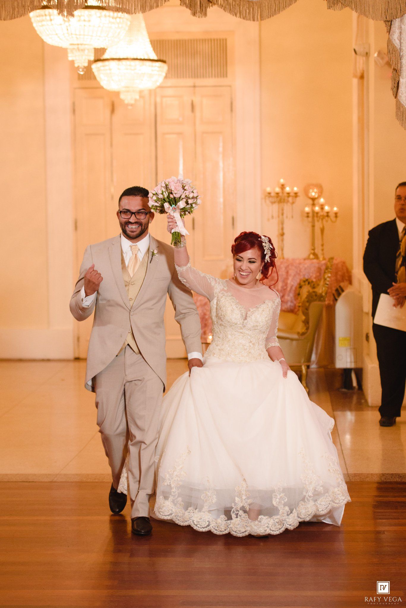 Joyful newlyweds make grand entrance, bride in lace gown holding bouquet, groom in tan suit at wedding reception.