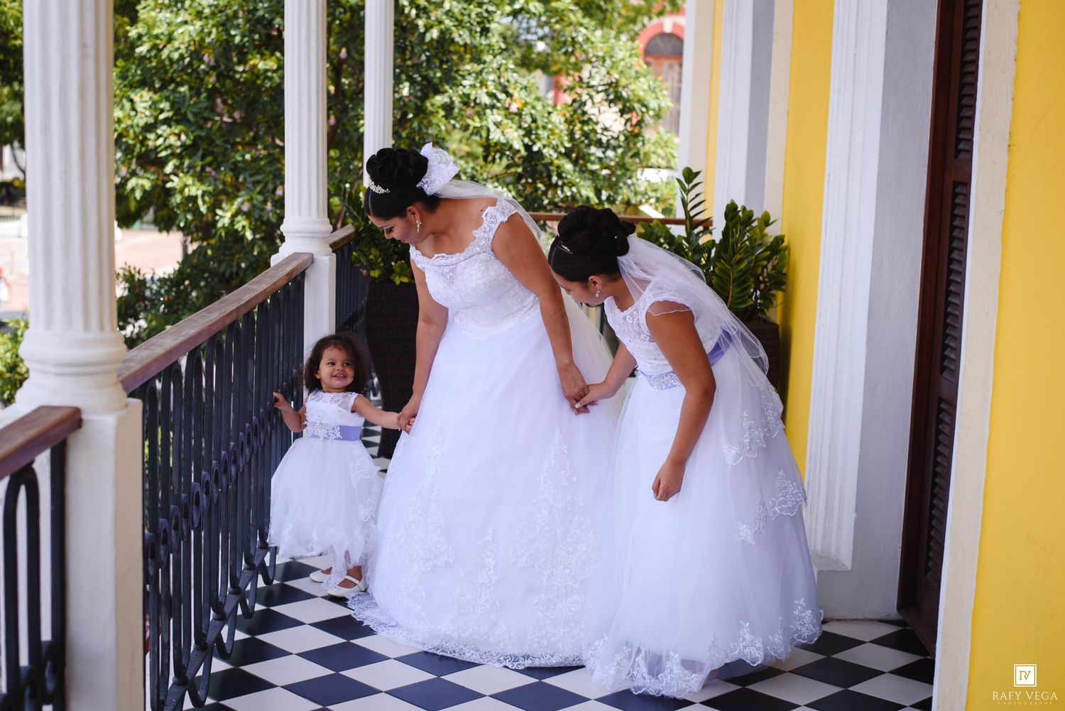 Bride and flower girls in white dresses on a checkered floor porch with columns.