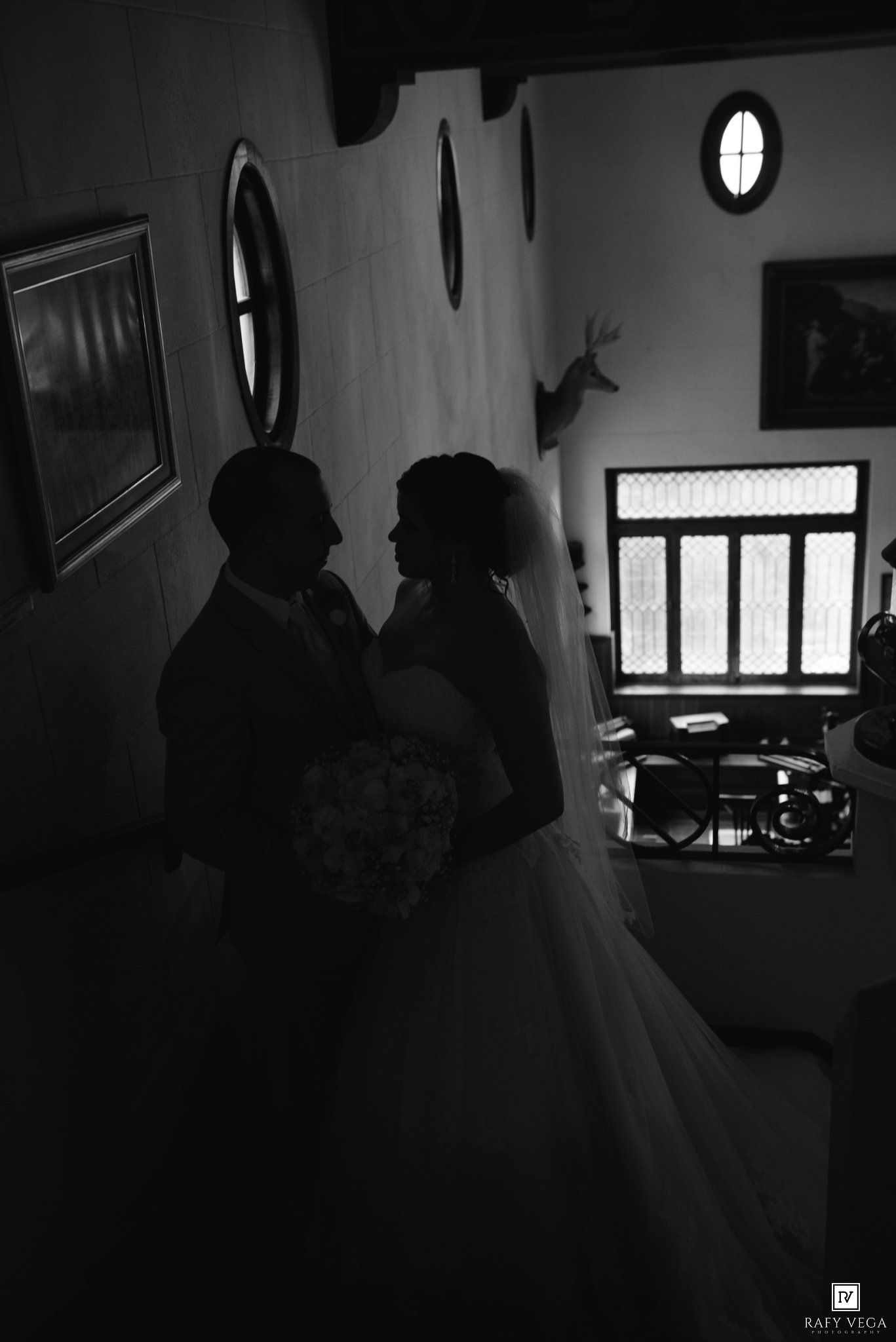 Black and white silhouette of a bride and groom embracing indoors, bride holding a bouquet, soft light from nearby window.