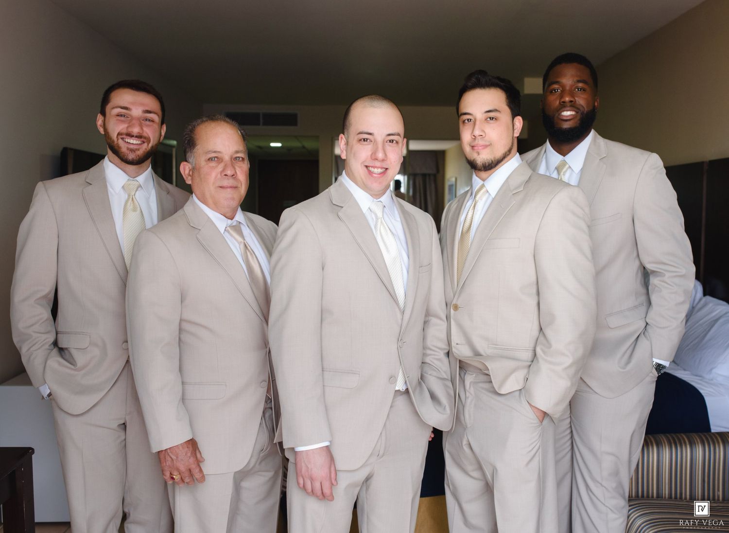 Five groomsmen in matching tan suits smiling together indoors before a wedding ceremony.