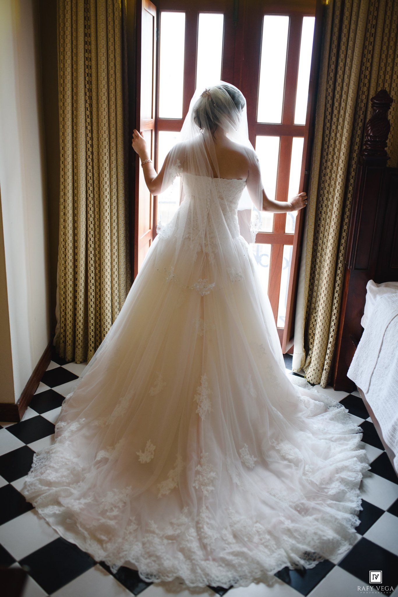 Bride in elegant white ball gown with lace train and veil standing at window on checkered floor.