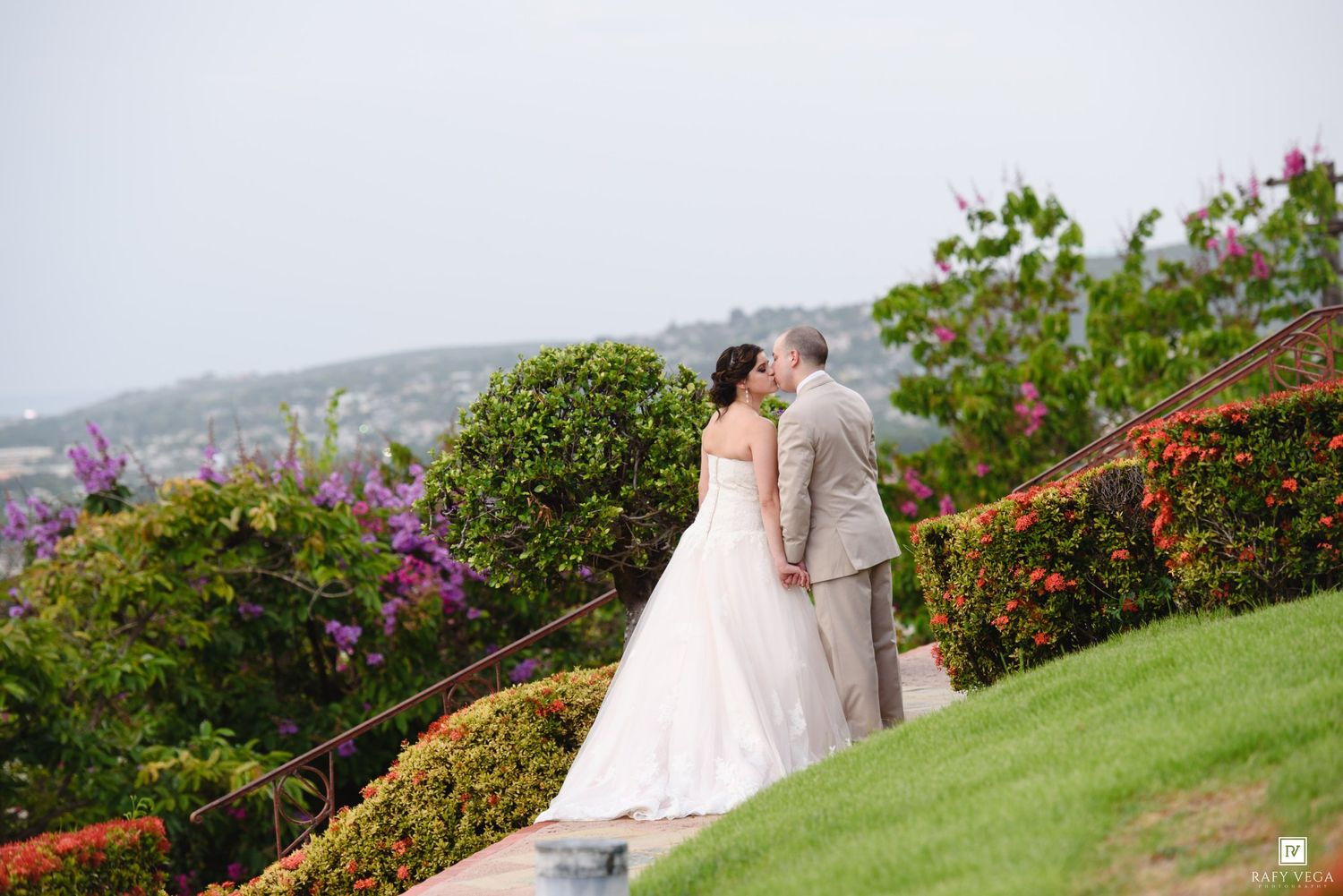 Bride and groom share a kiss at an outdoor wedding venue with lush greenery and purple flowers overlooking a coastal view.