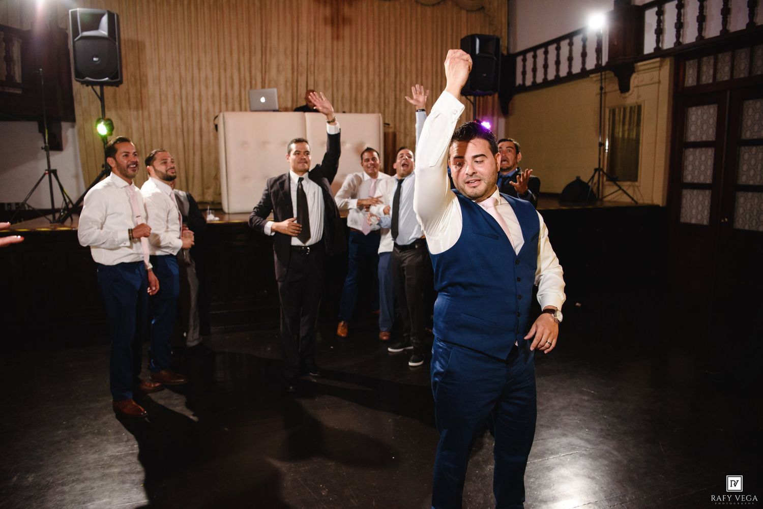 Groom in blue vest dancing with hands raised at wedding reception while groomsmen celebrate on dance floor.