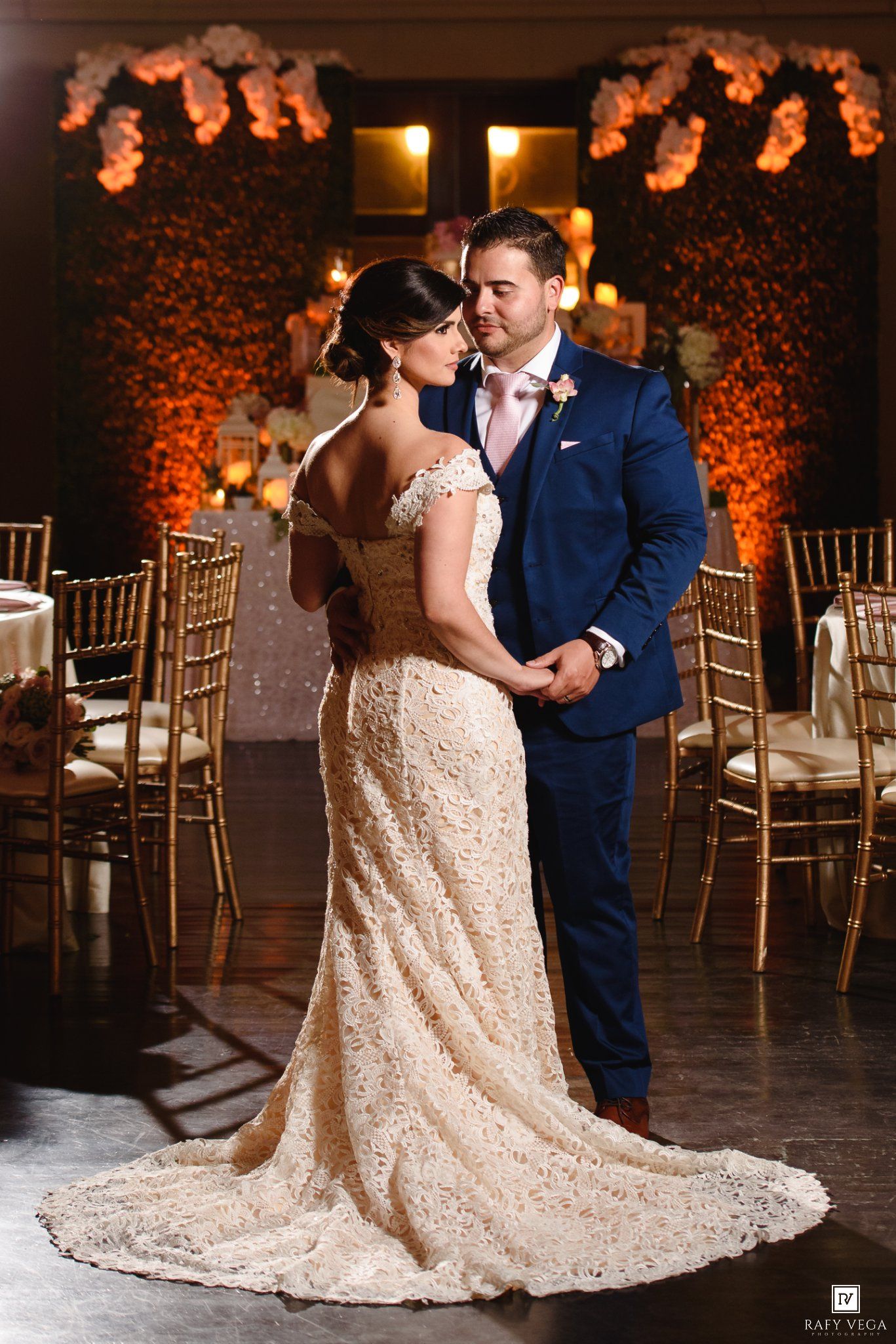 Bride in lace gown and groom in navy suit share romantic moment at elegantly decorated wedding reception venue.