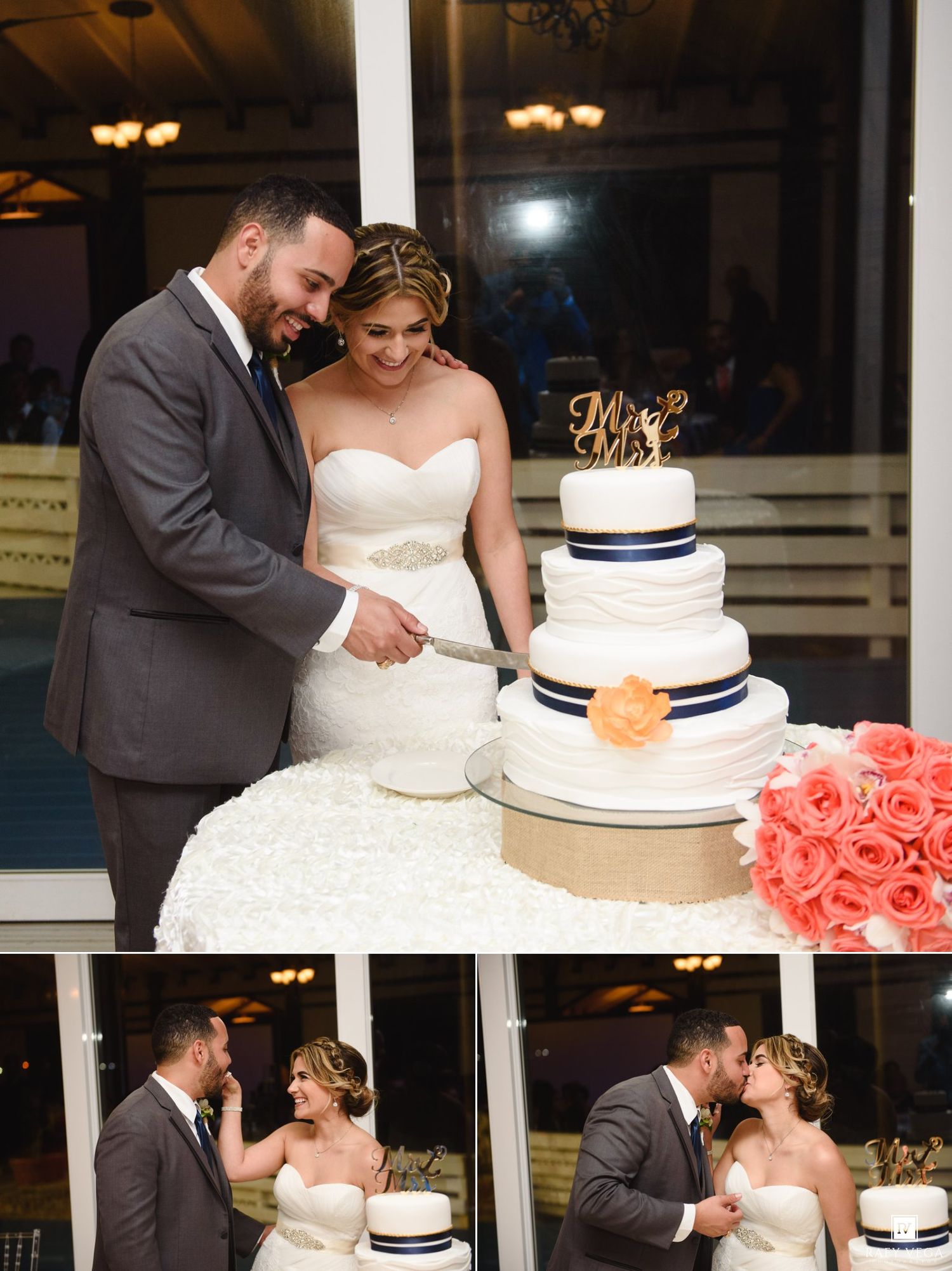 Bride and groom cutting their white three-tier wedding cake decorated with navy ribbon and coral roses at their reception.