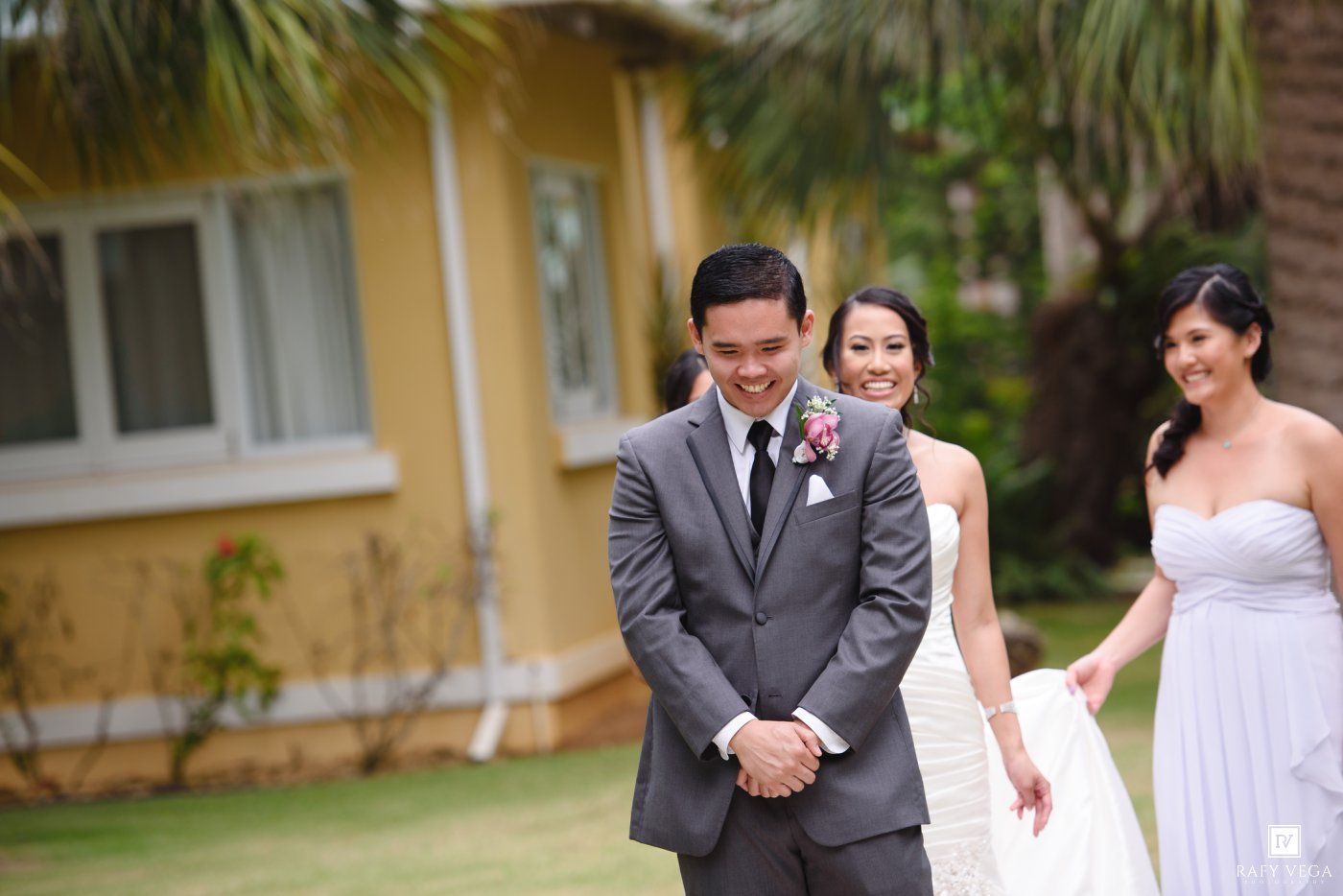 Groom in gray suit smiling as bride in white gown approaches with bridesmaid outdoors at wedding venue.