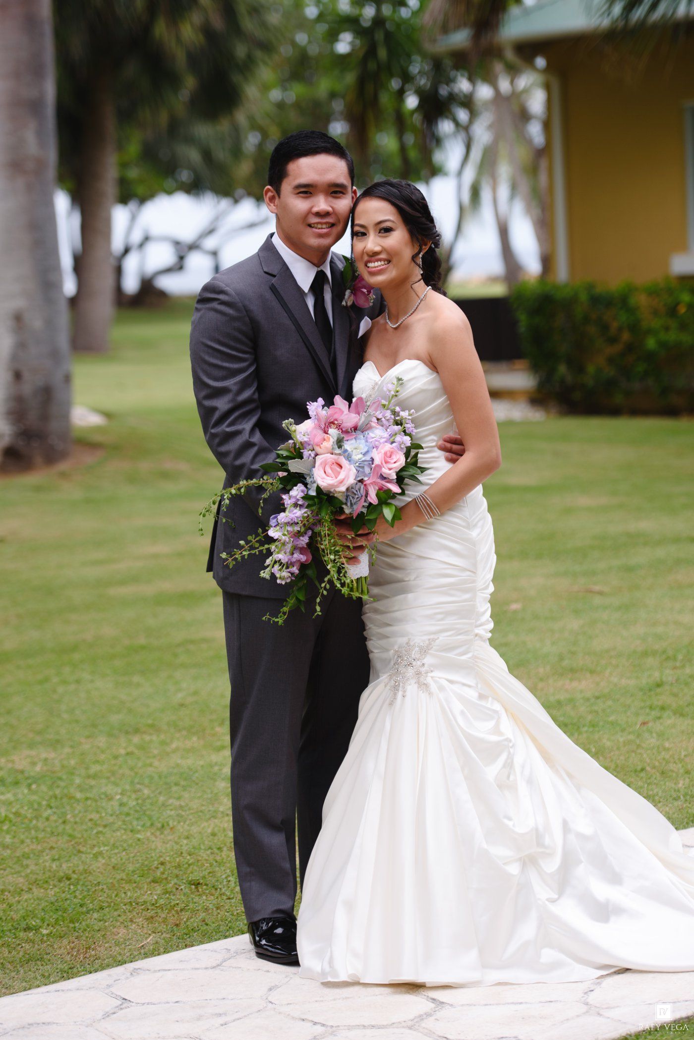 Bride and groom pose outdoors on wedding day, she holds pink bouquet in mermaid gown, he wears gray suit.