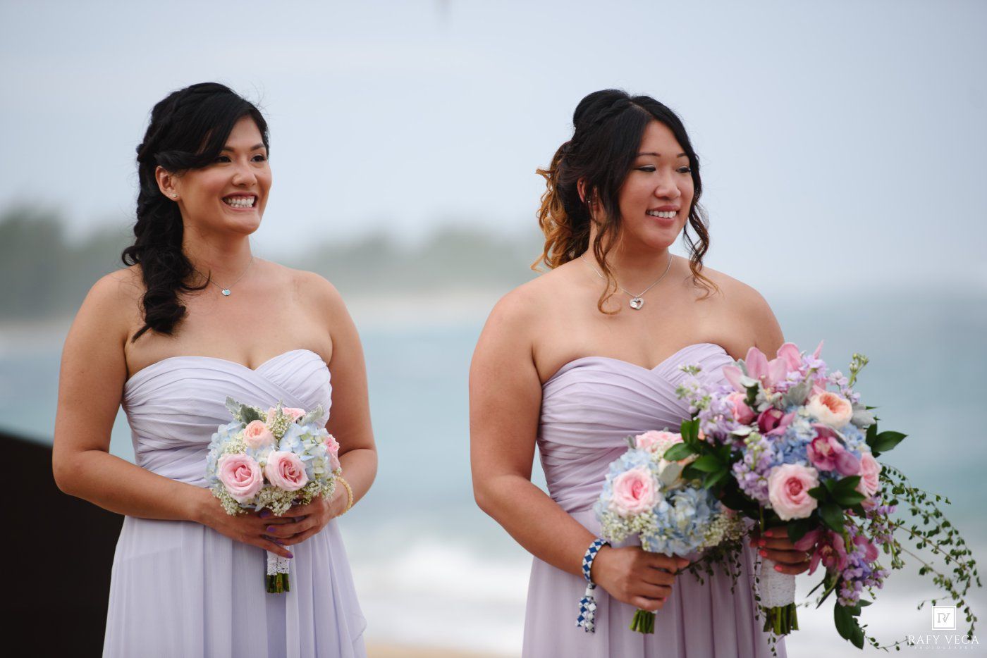 Two bridesmaids in lavender strapless gowns hold pink and blue bouquets at a beach wedding.