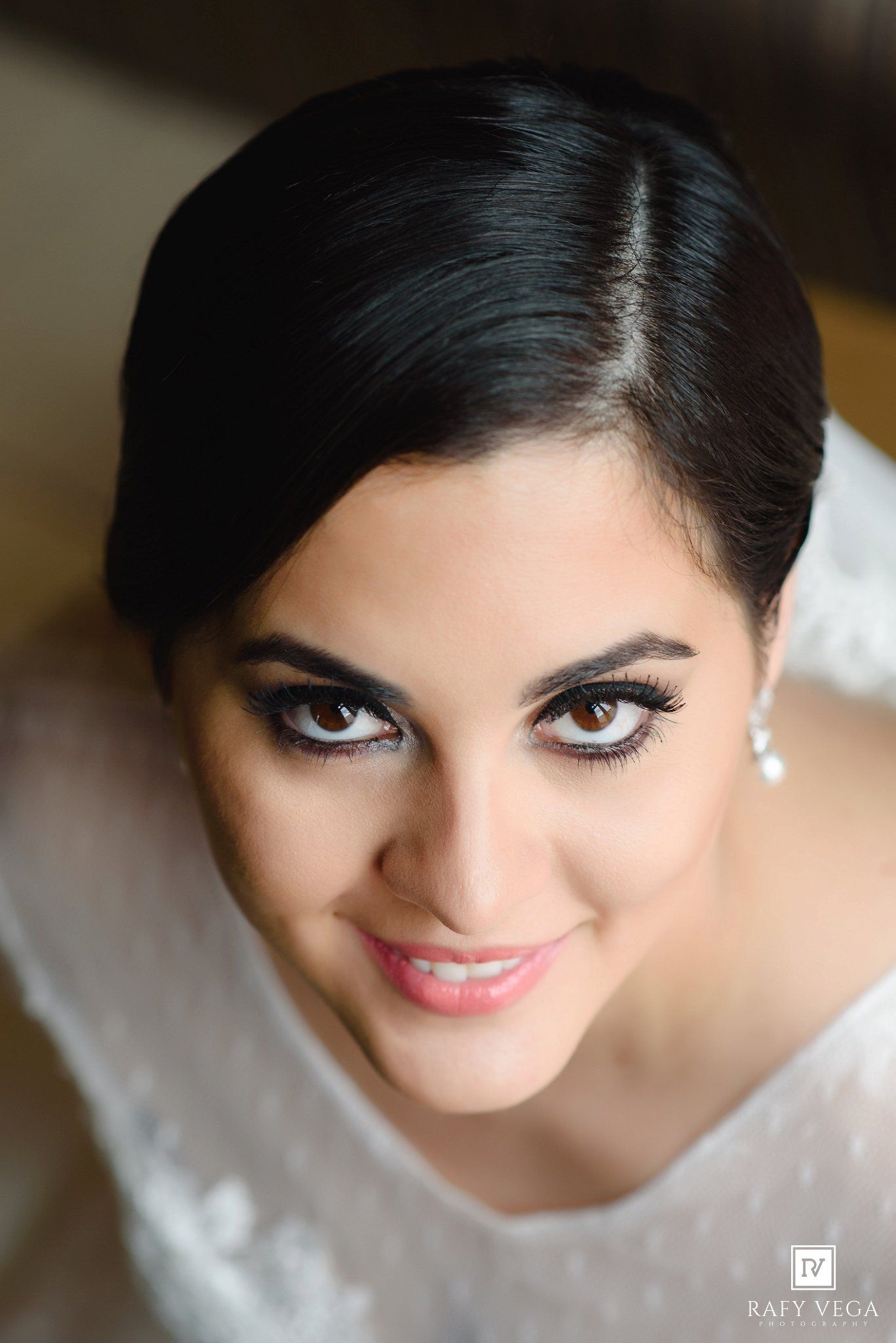 Elegant bride with sleek updo, dramatic eye makeup, and pearl earrings smiling in a white lace gown.