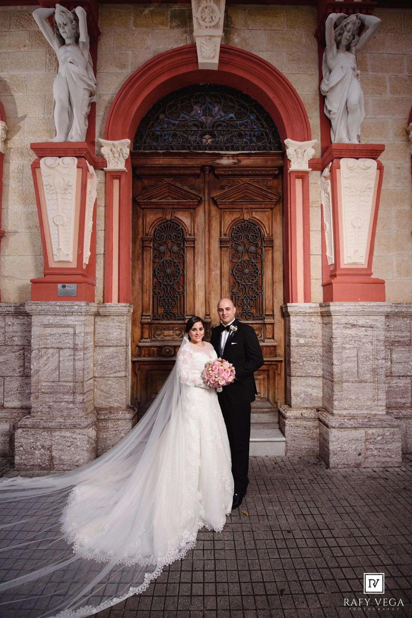 Bride and groom posing in front of ornate wooden doors with classical statues at their wedding.