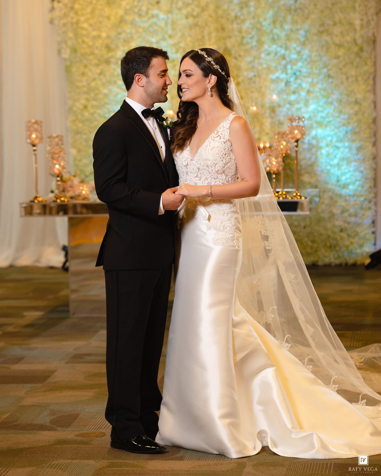 Bride and groom share a tender moment at elegant wedding reception with floral backdrop and candlelight.
