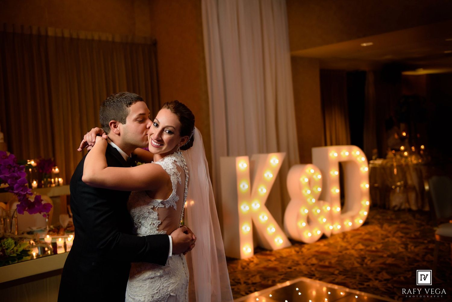 Bride and groom share first dance in front of illuminated marquee letters K&D at elegant wedding reception.