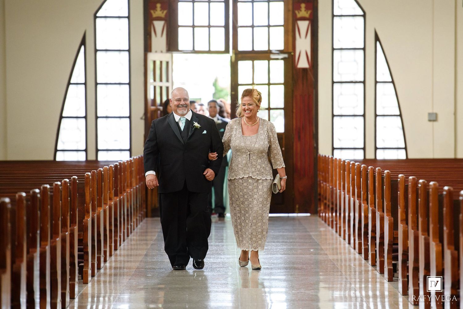 Elegant couple walking down church aisle during wedding ceremony, dressed in formal attire, with wooden pews and stained glass windows.