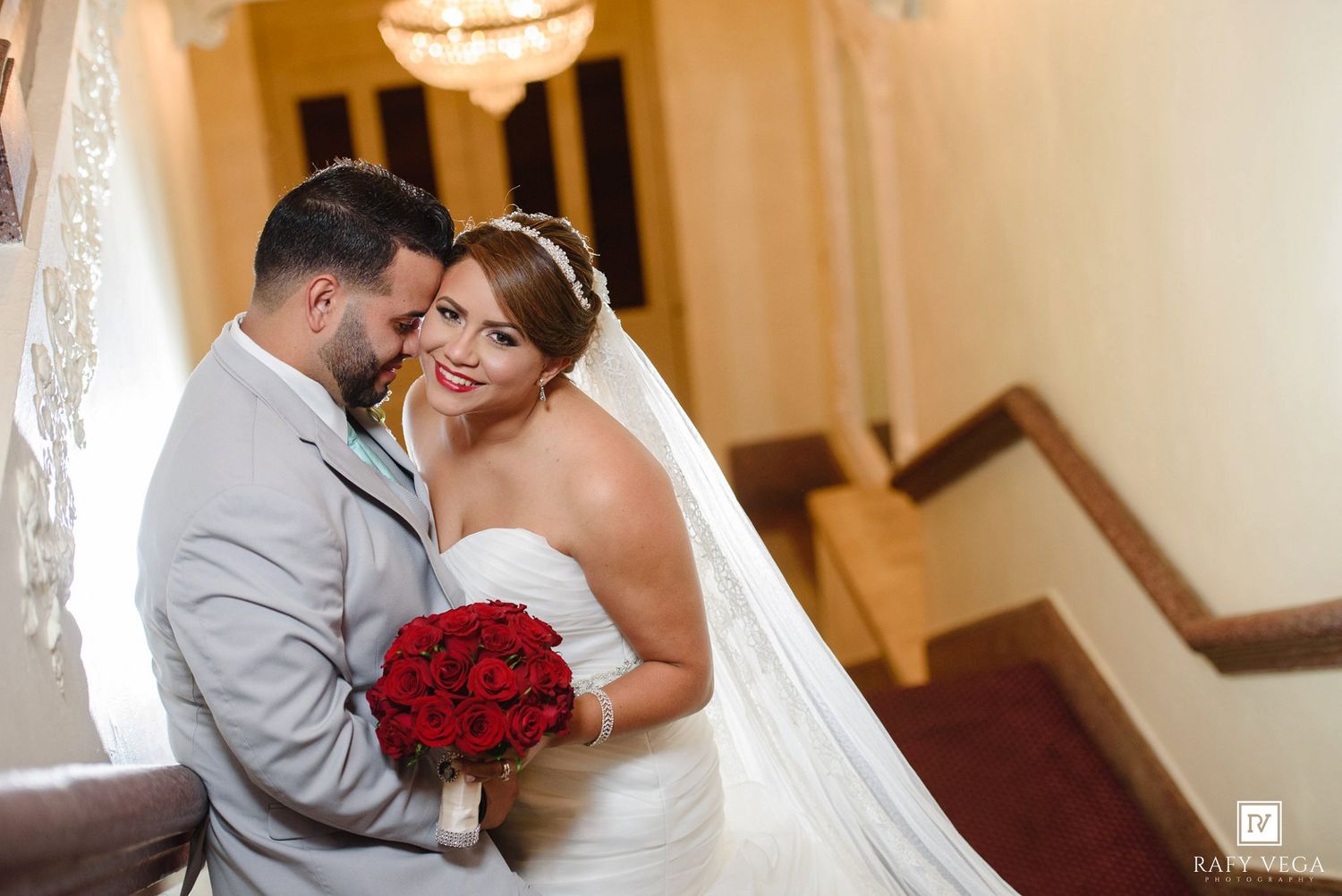 Bride in white gown holding red roses bouquet embraces groom in gray suit on elegant staircase.
