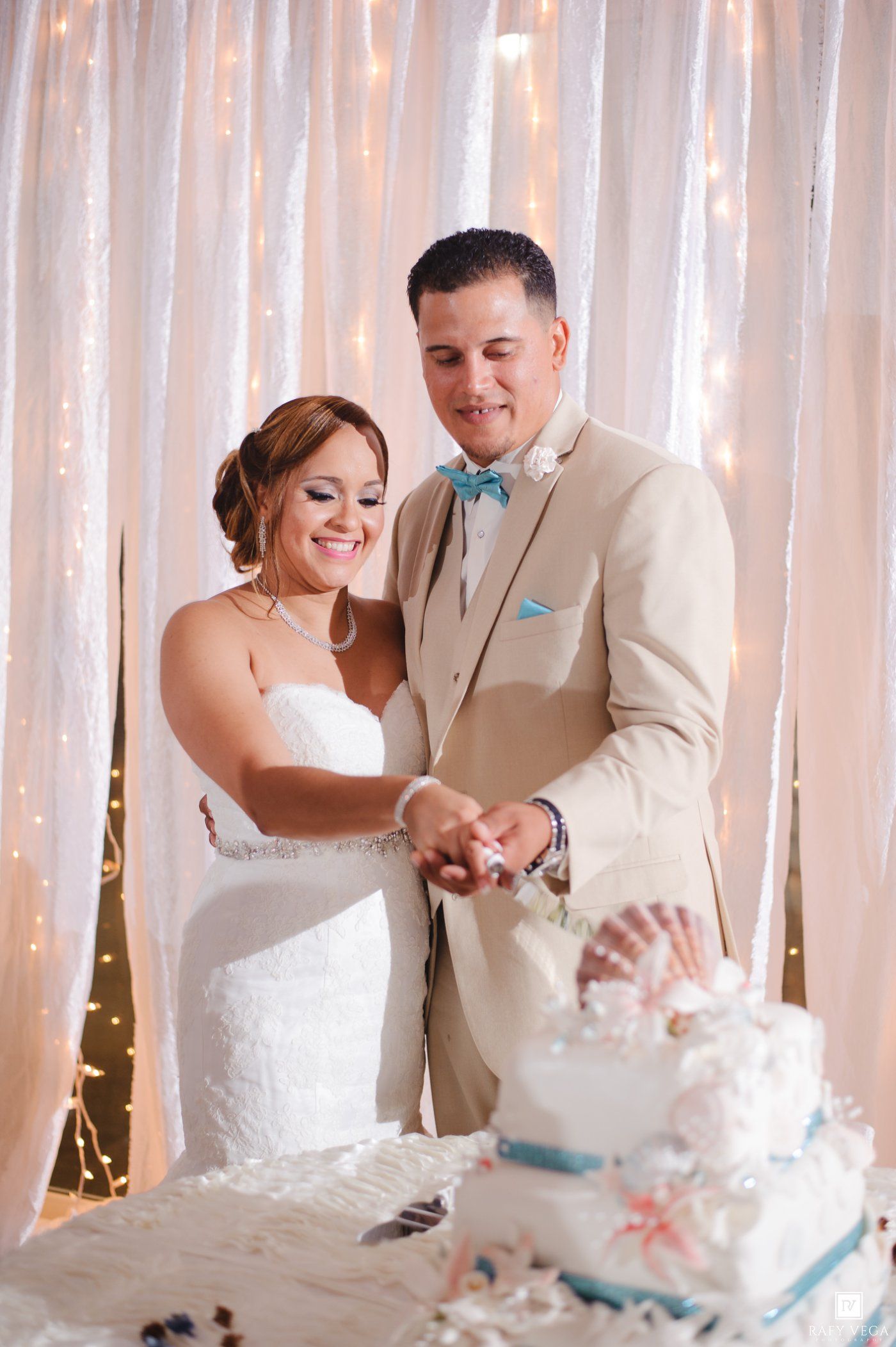 Bride and groom cutting their beach-themed white wedding cake together in front of a lit white drape backdrop.