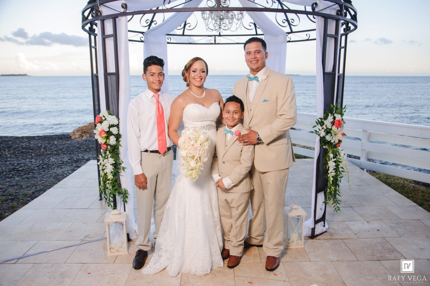 Happy wedding family portrait under coastal gazebo with bride, groom, and two boys in formal attire.