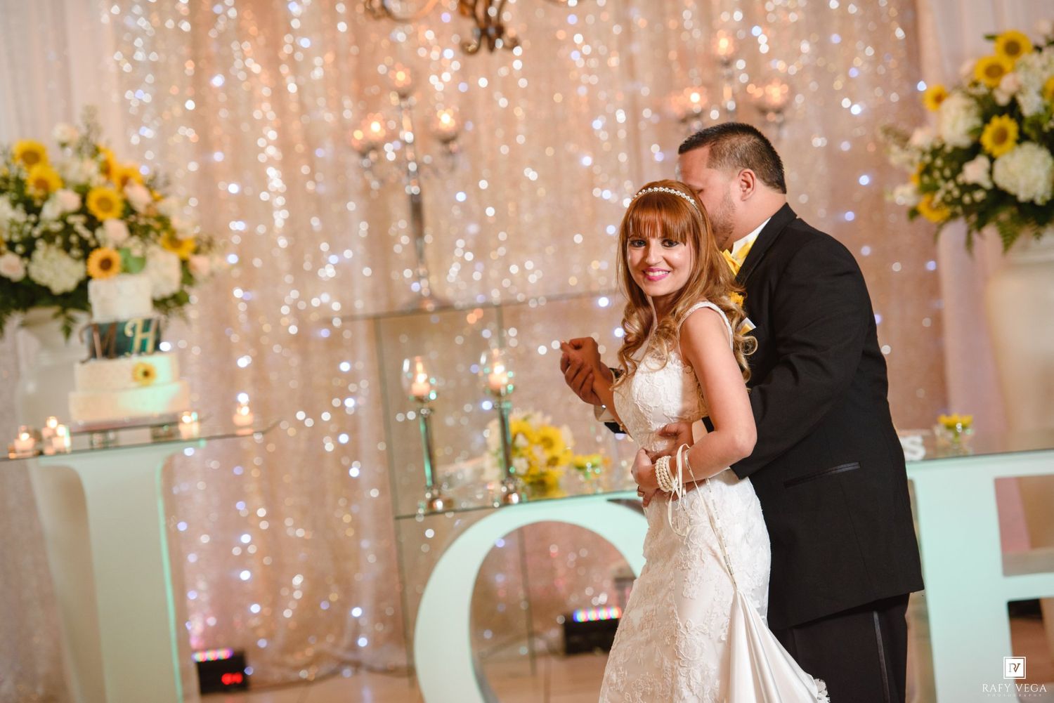 Bride and groom sharing first dance at elegant wedding reception with sparkling light curtain backdrop and floral arrangements.