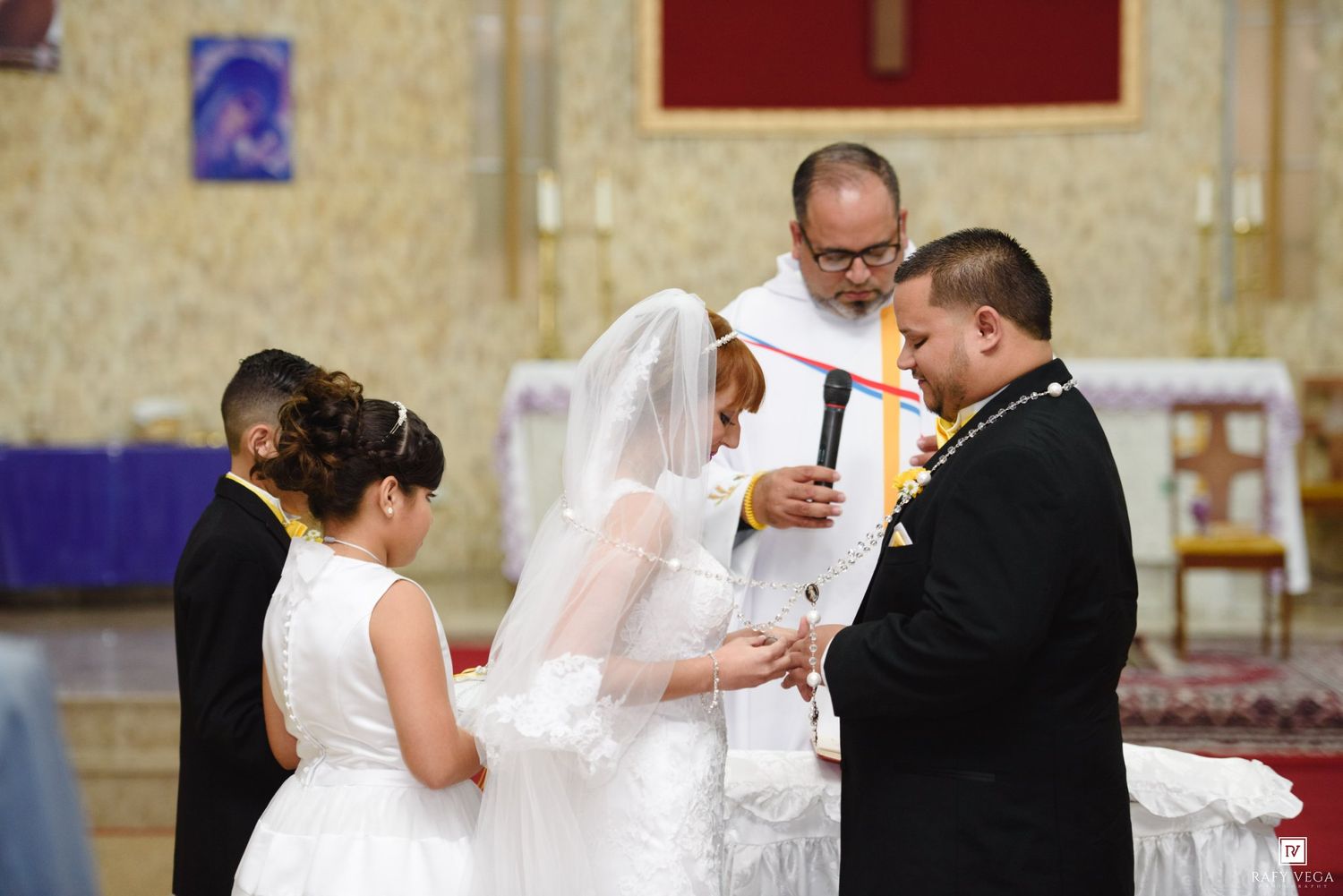 Bride and groom exchanging vows during Catholic church wedding ceremony with officiant and children present.