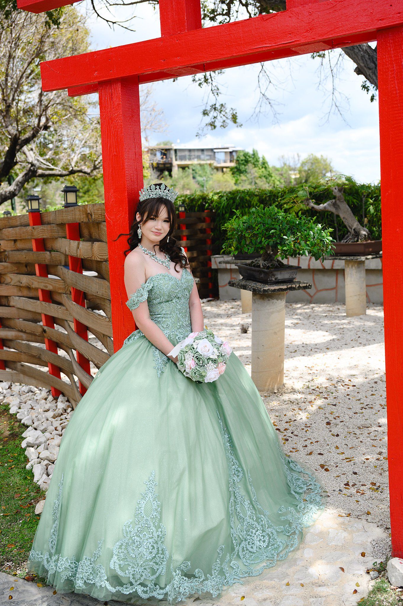 Young woman in sage green quinceañera ball gown with tiara holds bouquet under red torii gate in garden.