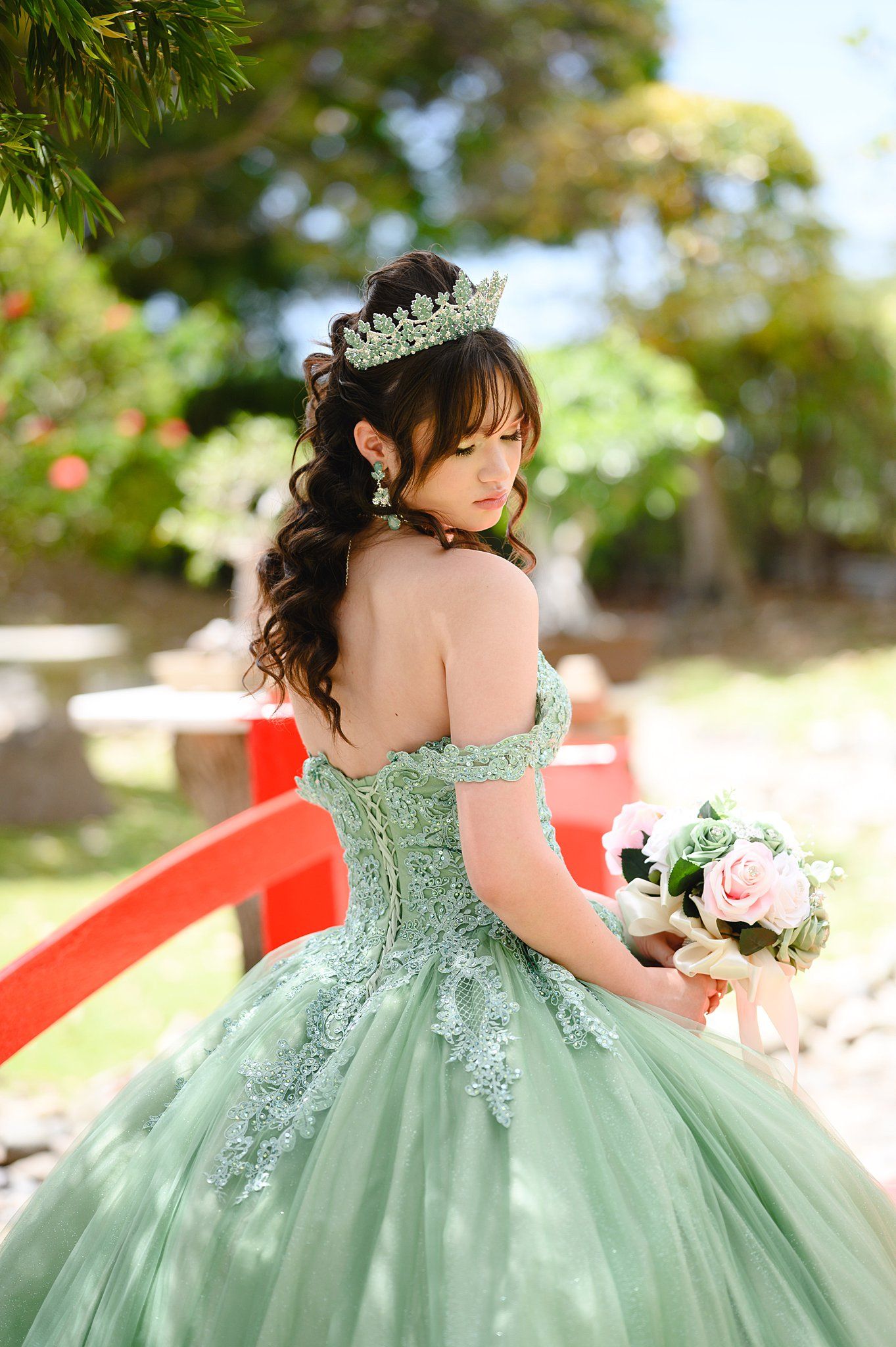 Young woman in sage green off-shoulder ballgown with tiara holds pink bouquet in lush garden setting.